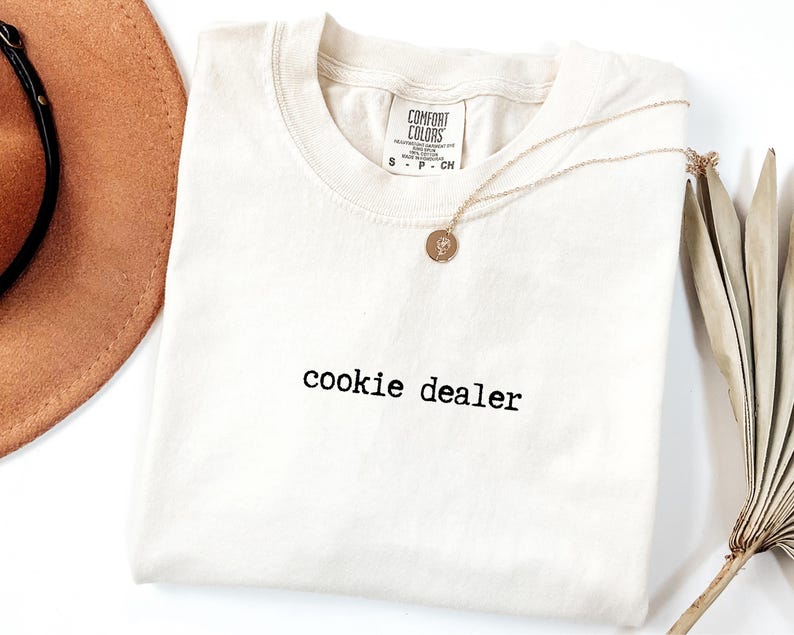 Pu&ograve; includere: T-shirt color crema con la scritta "cookie dealer" stampata in nero. Una collana d'oro con un ciondolo circolare &egrave; appoggiata sulla maglietta. Un cappello marrone e steli di piante secche sullo sfondo.