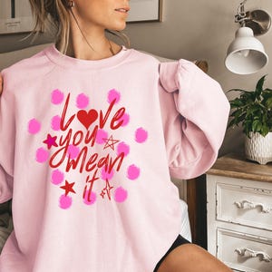 May include: A light pink sweatshirt with the phrase "Love You Mean It" in red script, a red heart, pink stars, and pink polka dots. The sweatshirt is a long-sleeved crew neck.