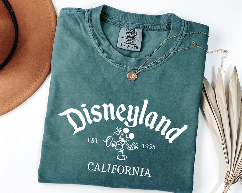 Może przedstawiać: Niebiesko-zielona koszulka z napisem "Disneyland" w białym skrypcie, wraz z "Est. 1955" i "California". Złoty naszyjnik z wisiorkiem spoczywa na koszulce. Widoczny jest r&oacute;wnież brązowy kapelusz i suszone liście.