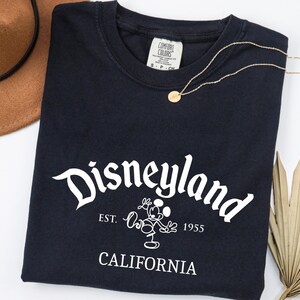 Może przedstawiać: Granatowa koszulka z napisem "Disneyland" w białym skrypcie, wizerunkiem Myszki Miki i tekstem "EST. 1955 CALIFORNIA". Widoczny jest r&oacute;wnież złoty naszyjnik i brązowy kapelusz.