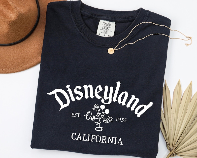 Może przedstawiać: Granatowa koszulka z napisem "Disneyland" w białym skrypcie, wizerunkiem Myszki Miki i tekstem "EST. 1955 CALIFORNIA". Widoczny jest r&oacute;wnież złoty naszyjnik i brązowy kapelusz.