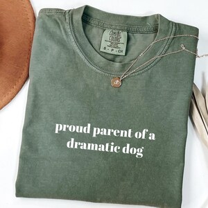 May include: An olive green t-shirt with the phrase "proud parent of a dramatic dog" in white lettering. A gold necklace with a pendant rests on the shirt. A brown hat and dried plant are also visible.