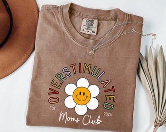 May include: A brown Comfort Colors t-shirt with the words "OVERSTIMULATED Moms Club" in a rainbow arc above a smiling daisy graphic. The shirt also has the text "EST. 2025". A silver necklace is draped across the shirt.