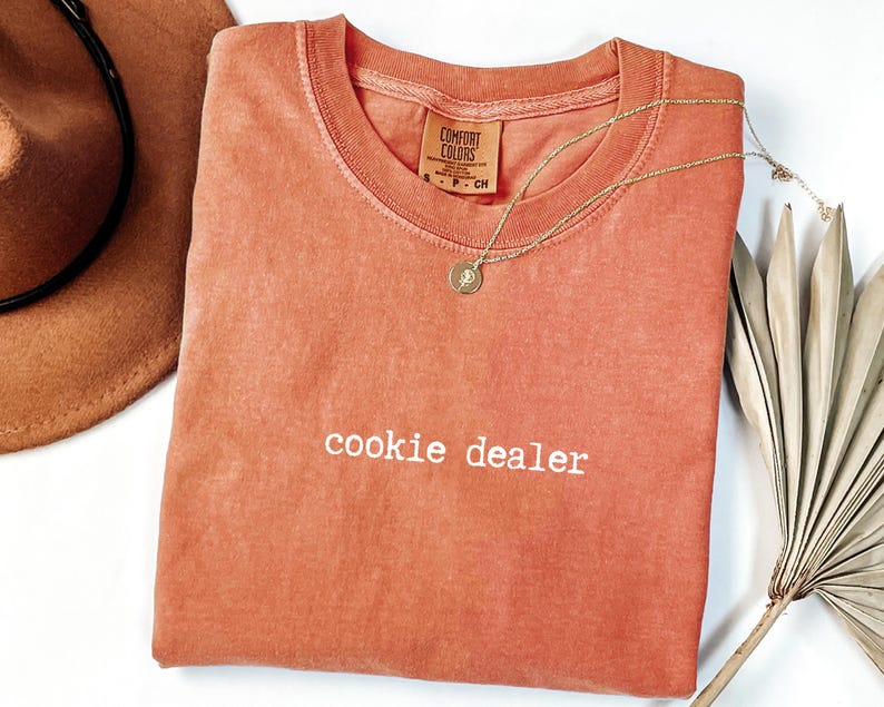 Pu&ograve; includere: Una maglietta Comfort Colors color corallo con la scritta "cookie dealer" in bianco. Una collana d'oro con un ciondolo poggia sulla maglietta. Un cappello marrone e una foglia di palma secca sono anch'essi nell'immagine.