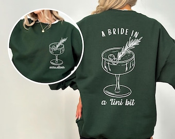 May include: Dark green sweatshirt with a white graphic of a cocktail and the text "A Bride in a tini bit". The front of the sweatshirt has a smaller cocktail graphic and the text "mrs.oliver".