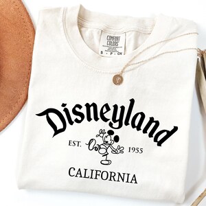Może przedstawiać: Kremowa koszulka z napisem "Disneyland" w czarnym, stylizowanym kroju pisma. Poniżej znajduje się grafika Myszki Miki i tekst "EST. 1955 CALIFORNIA". Widoczny jest złoty naszyjnik i brązowy kapelusz.