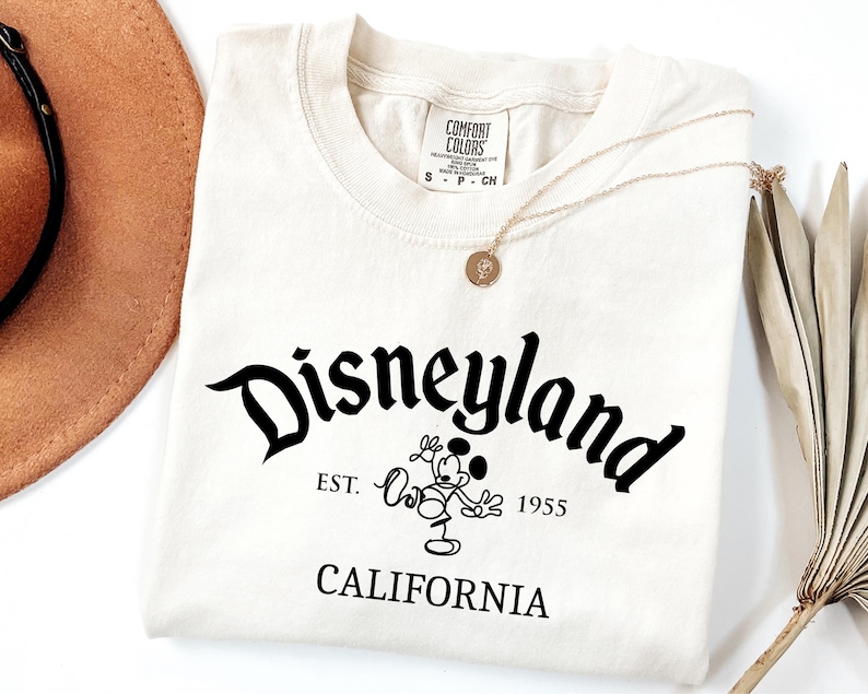 Może przedstawiać: Kremowa koszulka z napisem "Disneyland" w czarnym, stylizowanym kroju pisma. Poniżej znajduje się grafika Myszki Miki i tekst "EST. 1955 CALIFORNIA". Widoczny jest złoty naszyjnik i brązowy kapelusz.