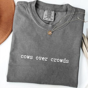 May include: A gray Comfort Colors t-shirt with the phrase "cows over crowds" in white lettering. A gold pendant necklace and a brown hat are also visible. The shirt is folded and lies on a white surface.