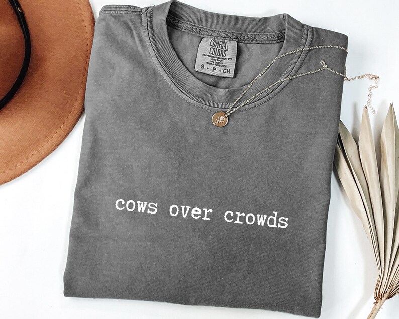 May include: A gray Comfort Colors t-shirt with the phrase "cows over crowds" in white lettering. A gold pendant necklace and a brown hat are also visible. The shirt is folded and lies on a white surface.