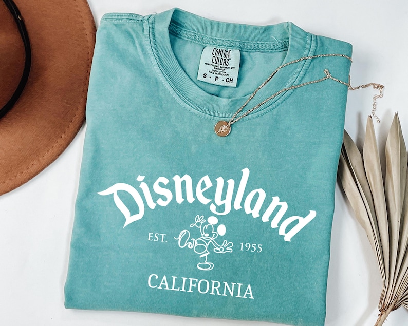 Może przedstawiać: Turkusowa koszulka Comfort Colors z napisem "Disneyland" w białym skrypcie, grafiką Myszki Miki i tekstem "Est. 1955 California". Złoty naszyjnik z wisiorkiem spoczywa na koszulce.