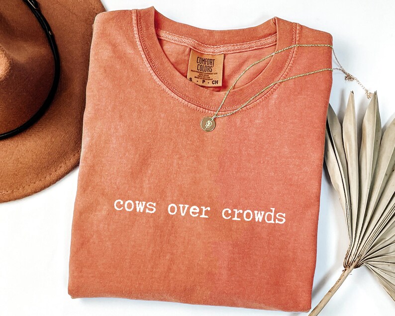 May include: A coral-colored Comfort Colors t-shirt with the phrase "cows over crowds" in white lettering. A gold necklace with a pendant rests on the shirt. A brown cowboy hat and dried palm frond are also in the image.