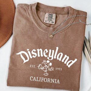 Może przedstawiać: Brązowa koszulka z białym napisem "Disneyland", postacią Myszki Miki i tekstem "Est. 1955 California". Srebrny naszyjnik z wisiorkiem leży na koszulce. Brązowy kapelusz i suszone liście w tle.