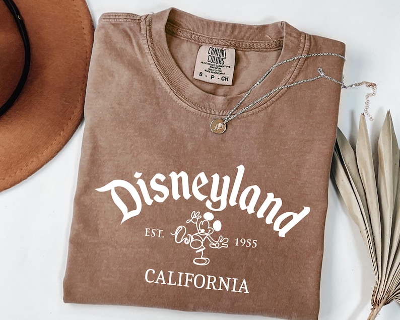 Może przedstawiać: Brązowa koszulka z białym napisem "Disneyland", postacią Myszki Miki i tekstem "Est. 1955 California". Srebrny naszyjnik z wisiorkiem leży na koszulce. Brązowy kapelusz i suszone liście w tle.
