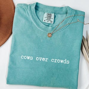 May include: A turquoise Comfort Colors t-shirt with the phrase "cows over crowds" in white lettering. A gold necklace with a pendant rests on the shirt. A brown hat and dried plant are in the background.