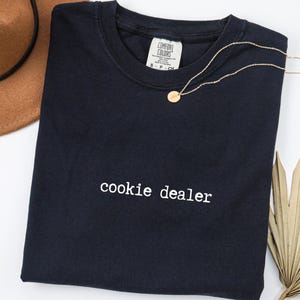 Pu&ograve; includere: Una maglietta blu navy con la scritta "cookie dealer" in bianco. Una collana d'oro con un piccolo ciondolo &egrave; appoggiata sulla maglietta. Un cappello marrone &egrave; visibile nell'angolo in alto a sinistra e una foglia di palma secca in basso a destra.