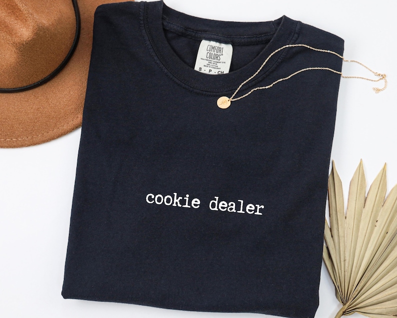 Pu&ograve; includere: Una maglietta blu navy con la scritta "cookie dealer" in bianco. Una collana d'oro con un piccolo ciondolo &egrave; appoggiata sulla maglietta. Un cappello marrone &egrave; visibile nell'angolo in alto a sinistra e una foglia di palma secca in basso a destra.
