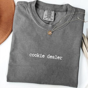 Pu&ograve; includere: Una maglietta grigia Comfort Colors con la scritta "cookie dealer" in bianco. Una collana color oro con un piccolo ciondolo &egrave; appoggiata sulla maglietta. Un cappello marrone e una pianta secca sono sullo sfondo.