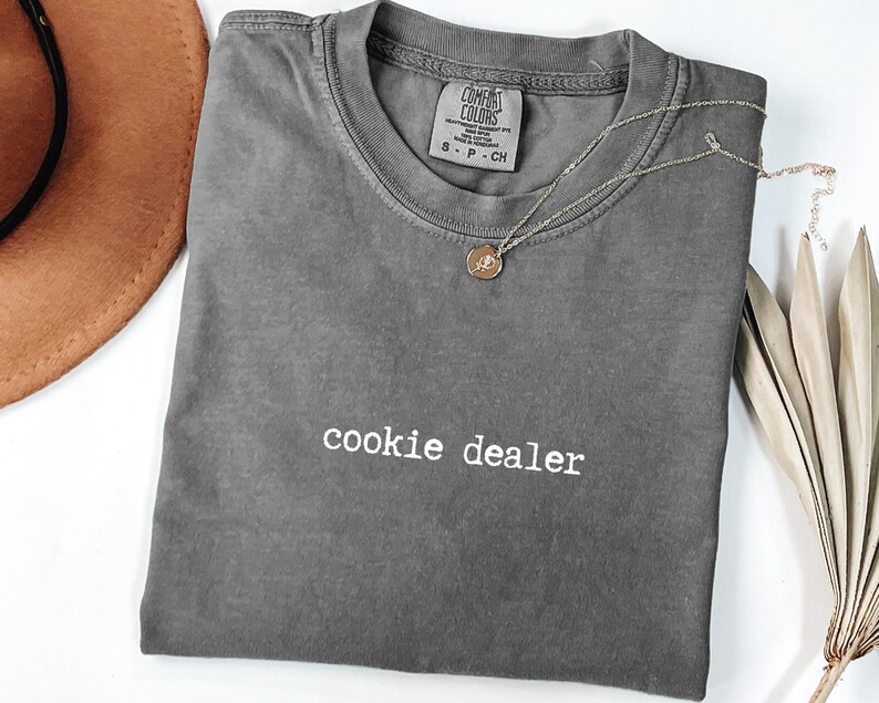 Pu&ograve; includere: Una maglietta grigia Comfort Colors con la scritta "cookie dealer" in bianco. Una collana color oro con un piccolo ciondolo &egrave; appoggiata sulla maglietta. Un cappello marrone e una pianta secca sono sullo sfondo.