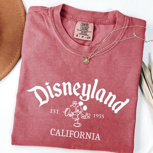 Może przedstawiać: Czerwona koszulka z napisem "Disneyland" w białym skrypcie, z grafiką Myszki Miki i napisem "California" poniżej. Złoty naszyjnik z wisiorkiem spoczywa na koszulce. Częściowo widoczny brązowy kapelusz.