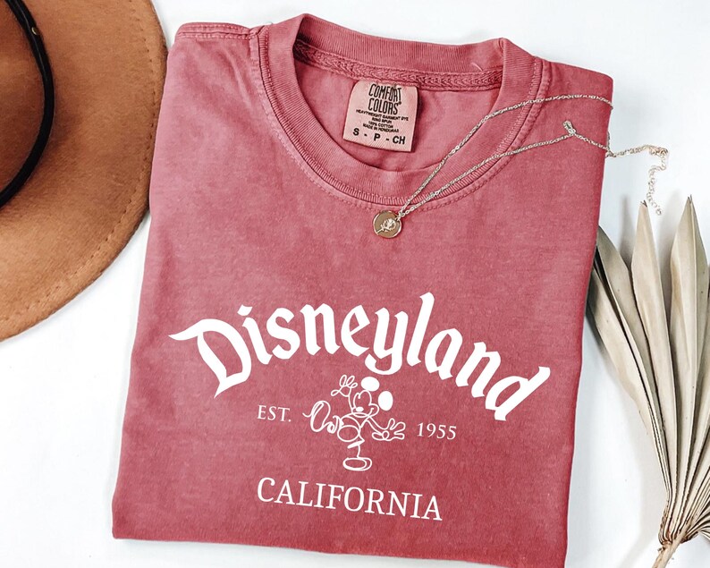 Może przedstawiać: Czerwona koszulka z napisem "Disneyland" w białym skrypcie, z grafiką Myszki Miki i napisem "California" poniżej. Złoty naszyjnik z wisiorkiem spoczywa na koszulce. Częściowo widoczny brązowy kapelusz.