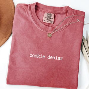 Pu&ograve; includere: Una maglietta colorata di rosso con la scritta "cookie dealer" stampata in bianco. Una collana d'oro con un ciondolo &egrave; appoggiata sulla maglietta. Un cappello marrone e una pianta secca sono sullo sfondo.