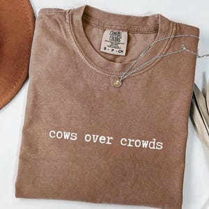 May include: A brown Comfort Colors t-shirt with the phrase "cows over crowds" in white lettering. A silver necklace with a small pendant rests on the shirt. A brown hat and dried plant stems are also visible.