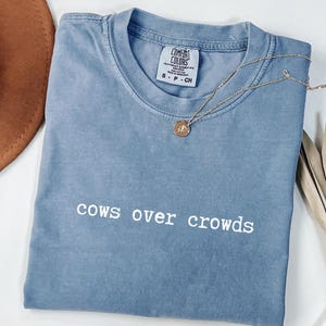 May include: A blue Comfort Colors t-shirt with the phrase "cows over crowds" in white. A gold necklace with a pendant rests on the shirt. A brown hat and dried palm leaves are in the background.
