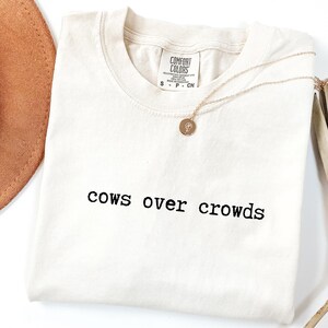 May include: Cream-colored t-shirt with the phrase "cows over crowds" printed in black. A gold necklace with a pendant and a brown hat are next to the shirt. The shirt has a Comfort Colors tag.