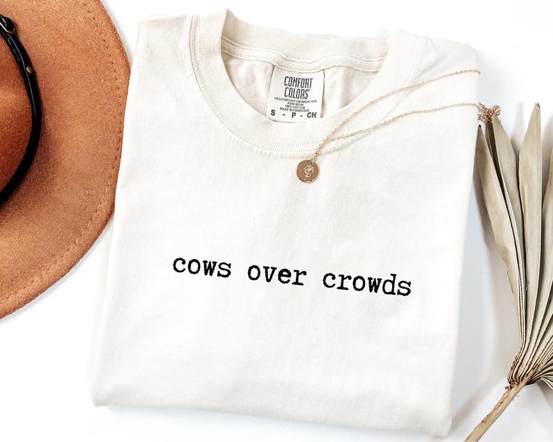 May include: Cream-colored t-shirt with the phrase "cows over crowds" printed in black. A gold necklace with a pendant and a brown hat are next to the shirt. The shirt has a Comfort Colors tag.