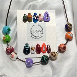 May include: A set of ten painted artificial nails with various designs, including a trident, gold accents, and geometric patterns. The nails are displayed on a white card with the text "Mage Manicures" and are accompanied by a beaded necklace.