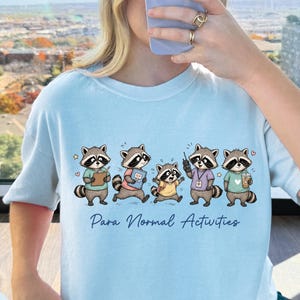 Puede incluir: Una camiseta azul claro con un gráfico de cinco mapaches de dibujos animados y el texto "Para Normal Activities" impreso en azul oscuro. Los mapaches aparecen en varias posturas, algunos sosteniendo objetos como un portapapeles, un libro y una bebida.
