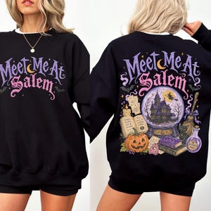 May include: A black crewneck sweatshirt featuring the text Meet Me At Salem in purple and pink on the front. The back displays a detailed graphic of a crystal ball with a house, a witch on a broom, a pumpkin, an open book, and a potion bottle.