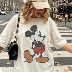 May include: Ivory t-shirt with a large Mickey Mouse graphic. The cartoon character is depicted in a classic pose. The shirt has short sleeves and a loose fit. The words "COMFORT COLORS" and "IVORY" are printed on the shirt.