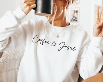 Puede incluir: Sudadera blanca de manga larga con el texto "Coffee & Jesus" en cursiva. Una persona sostiene una taza de café negra. La persona lleva un collar y hace el signo de la paz con los dedos.