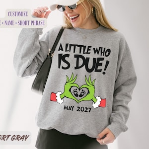 Puede incluir: Una sudadera gris deportivo de cuello redondo con el texto negro A LITTLE WHO IS DUE! sobre un gráfico de manos verdes formando un corazón con huellas de bebé. El diseño incluye puños rojos y el texto negro MAY 2027 en la parte inferior.