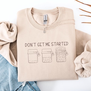 May include: A beige sweatshirt with the phrase "DON'T GET ME STARTED" printed in black. Below the text are three jar illustrations with tags. A silver necklace with a sun pendant is on the sweatshirt.