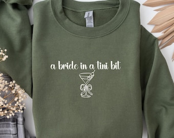 May include: An olive green sweatshirt with the phrase "a bride in a tini bit" in white script. A white line drawing of a martini glass with a bow is below the text. The sweatshirt is folded.