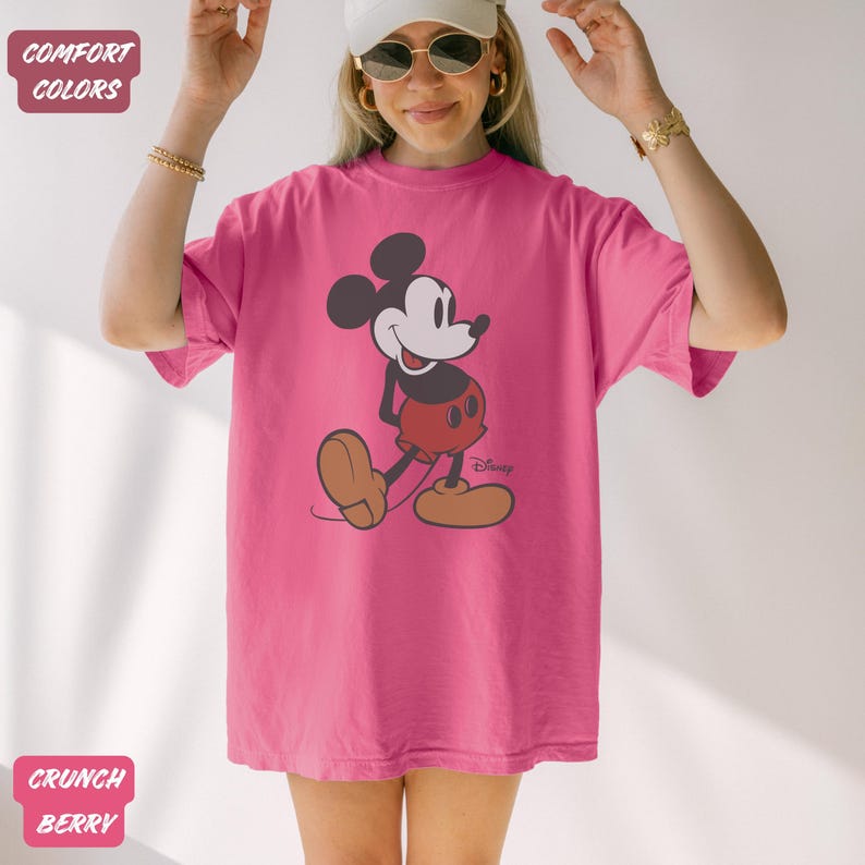May include: A magenta t-shirt with a Mickey Mouse graphic. The cartoon character is in black, red, and tan, with the Disney logo. The shirt has short sleeves and a loose fit. The words "Comfort Colors" and "Crunch Berry" are visible.