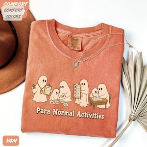 Peut inclure: Un t-shirt à col rond de couleur terre cuite orné d'un motif représentant cinq fantômes de dessin animé effectuant diverses tâches scolaires. Sous les fantômes, le texte "Para Normal Activities" est écrit dans une police brune arrondie.