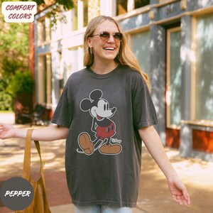May include: A dark grey t-shirt with a Mickey Mouse design. The cartoon mouse is wearing red shorts, brown shoes, and white gloves. The shirt has the word "Disney" printed below the character. The shirt is a comfort colors style.