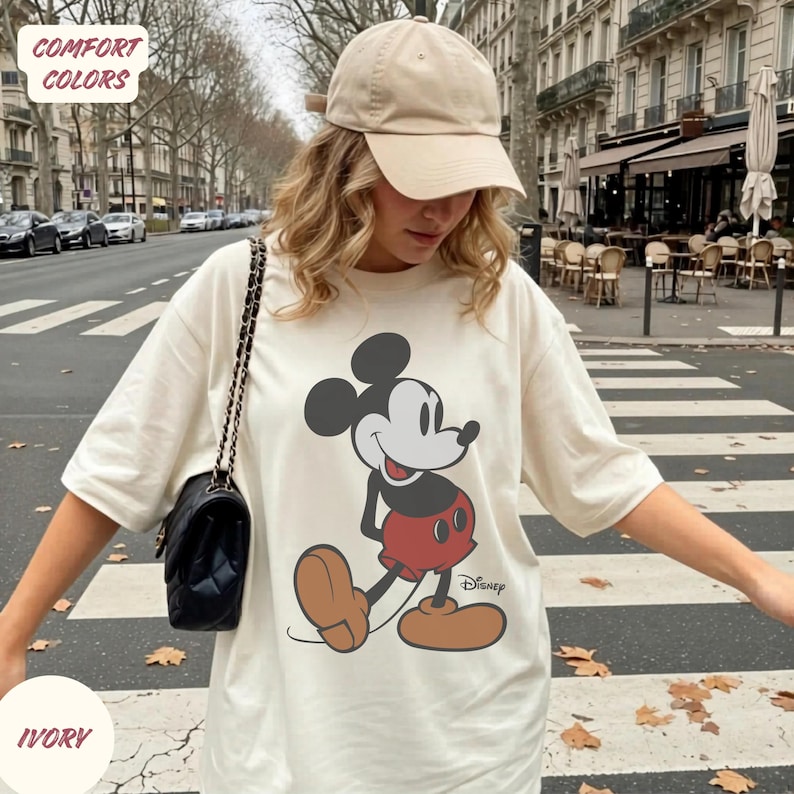 May include: Ivory t-shirt with a large Mickey Mouse graphic. The cartoon character is depicted in a classic pose. The shirt has short sleeves and a loose fit. The words "COMFORT COLORS" and "IVORY" are printed on the shirt.