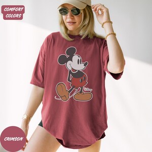 May include: Crimson t-shirt with a Mickey Mouse graphic. The cartoon character is shown in his classic outfit: black body, red shorts, and brown shoes. The shirt has a relaxed fit and short sleeves. The text "Disney" is below the graphic. The shirt is a comfort color.