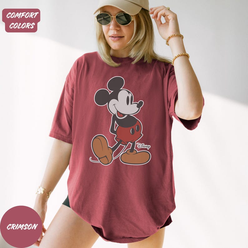 May include: Crimson t-shirt with a Mickey Mouse graphic. The cartoon character is shown in his classic outfit: black body, red shorts, and brown shoes. The shirt has a relaxed fit and short sleeves. The text "Disney" is below the graphic. The shirt is a comfort color.