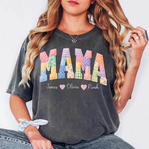 Peut inclure: T-shirt gris foncé avec le mot "MAMA" en lettres patchwork colorées. En dessous, les noms "James", "Olivia" et "Noah" avec des symboles de cœur. Le t-shirt est de coupe décontractée.