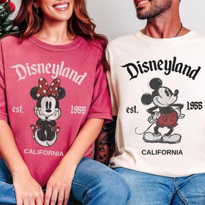 May include: Two t-shirts with Disneyland graphics. One is crimson with a Minnie Mouse design, the other is ivory with Mickey Mouse. Both shirts have "Disneyland est. 1955 California" printed on them.
