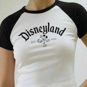 May include: White crop top with black raglan sleeves featuring the word "Disneyland" in a script font. Below the word is a graphic of Mickey Mouse and the text "EST. 1955". The shirt is paired with blue denim jeans.