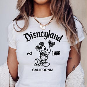 May include: White crop top featuring a black graphic of Mickey Mouse holding balloons, with the text "Disneyland est. 1955 CALIFORNIA". The shirt has short sleeves and a round neckline.