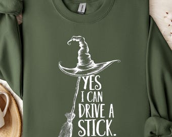 May include: An olive green sweatshirt featuring a white witch's hat and broom graphic, with the text "YES I CAN DRIVE A STICK". The garment is neatly folded on a white, fluffy surface. The sleeves are rolled up.