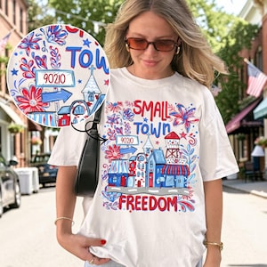 May include: White t-shirt with a colorful graphic design featuring the words "SMALL TOWN FREEDOM" and the number "90210". The design includes illustrations of buildings, flowers, and a water tower in red, white, and blue.