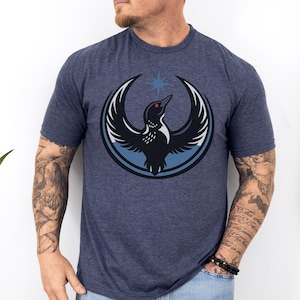 May include: Heather midnight navy t-shirt featuring a graphic of a bird with outstretched wings inside a blue circle. The bird is black with white accents and a red eye. A blue star is above the bird. The shirt is a crew neck.